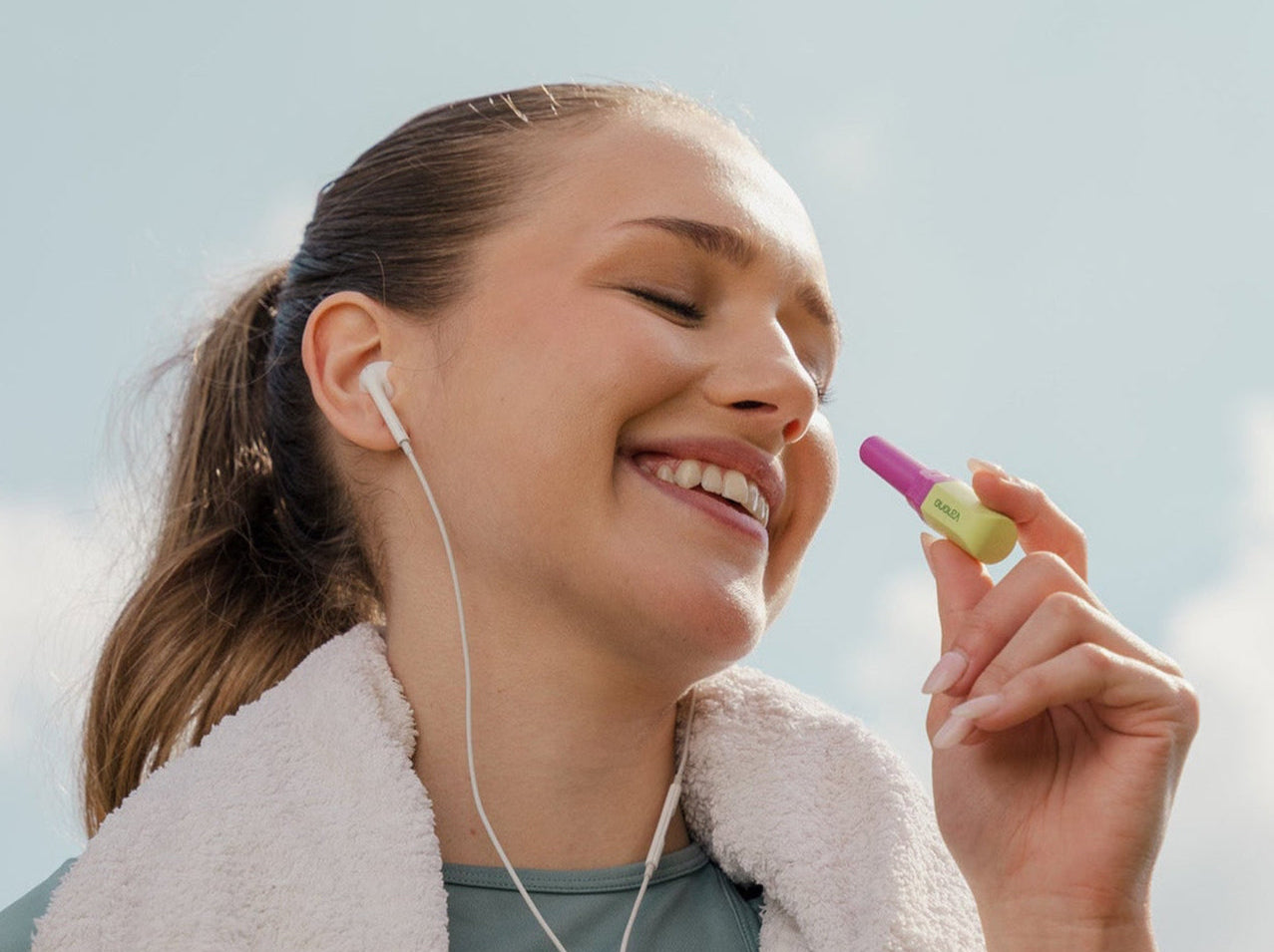 Woman outdoors with earphones, holding a small object, against a clear sky.